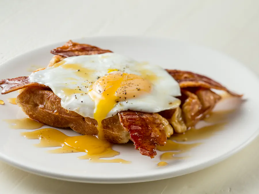 Liège waffle with a sunny side up egg and two strips of bacon, maple syrup being poured over the top